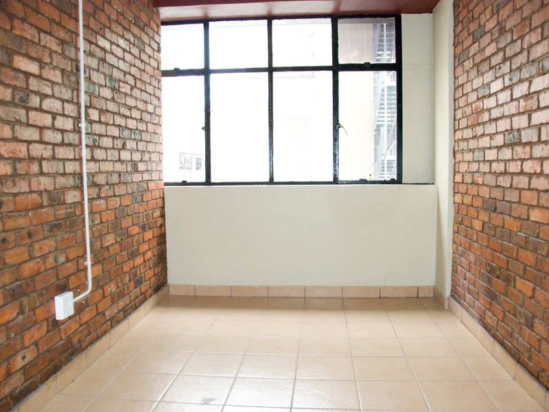 An empty bedroom featuring exposed brick walls, tiled flooring, and a large metal-framed window providing natural light.