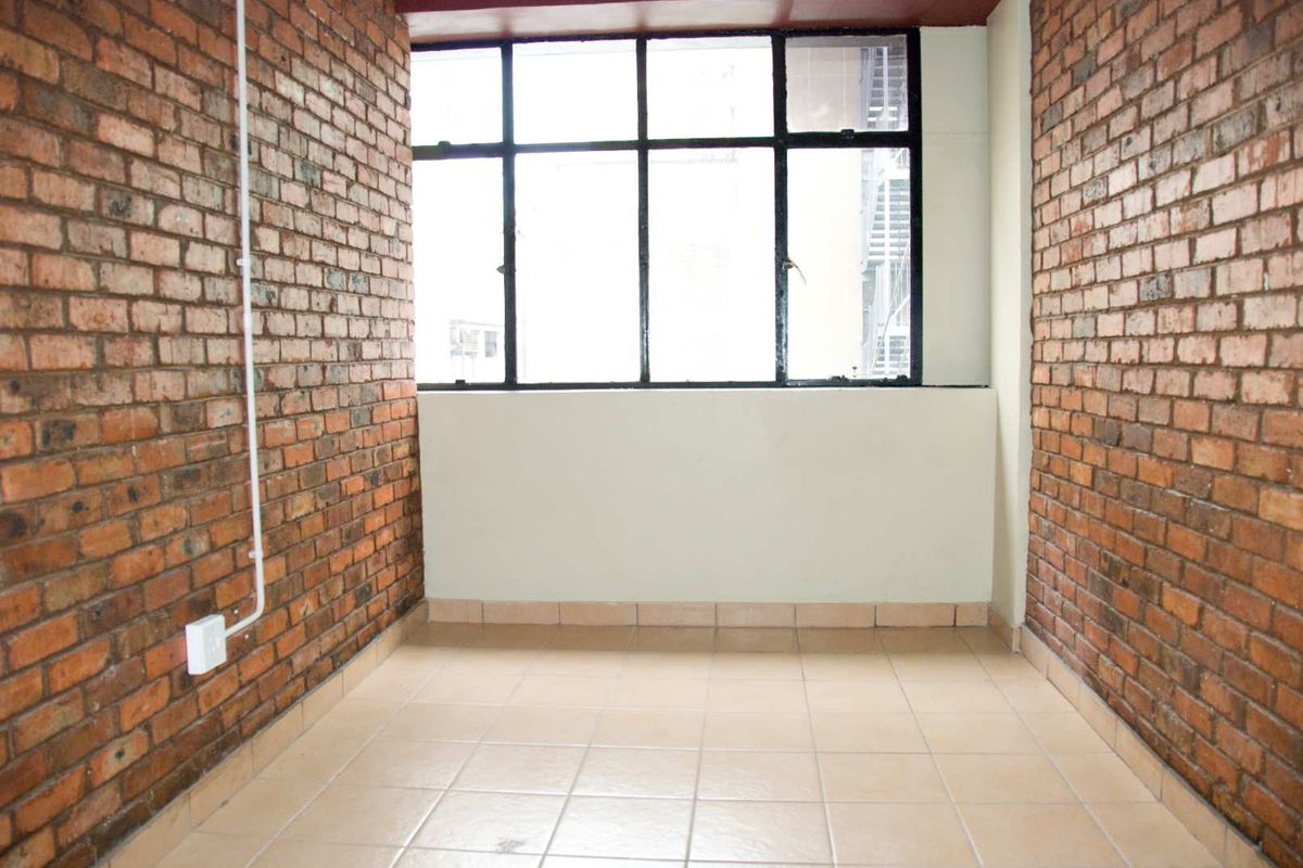 An empty bedroom featuring exposed brick walls, tiled flooring, and a large metal-framed window providing natural light.