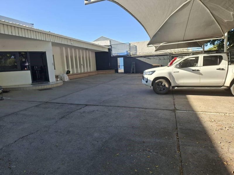 A paved rear exterior area featuring a large shade sail structure, concrete surfacing, and a secure metal gate entrance.