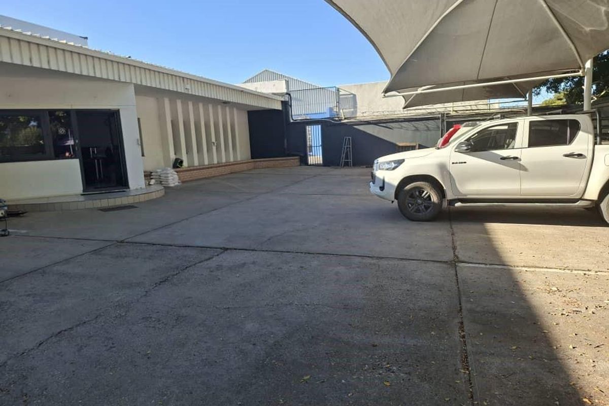 A paved rear exterior area featuring a large shade sail structure, concrete surfacing, and a secure metal gate entrance.