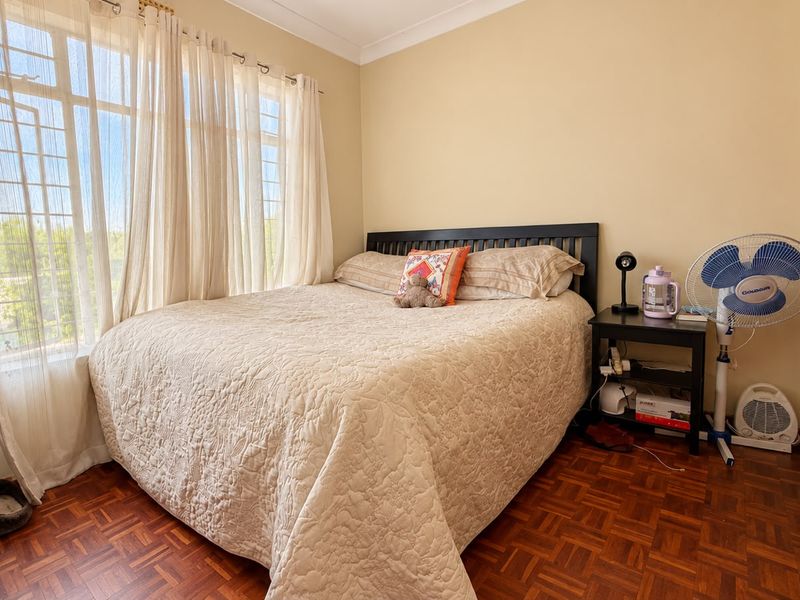 A cozy bedroom featuring classic parquet flooring, large windows with security bars, and neutral wall finishes, offering a comfortable and functional living space.