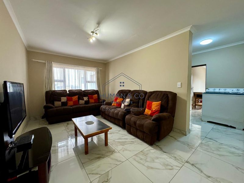 A well-maintained living room featuring polished marble-look floor tiles, neutral wall tones, and a bright, open layout connecting to the adjacent utility area.