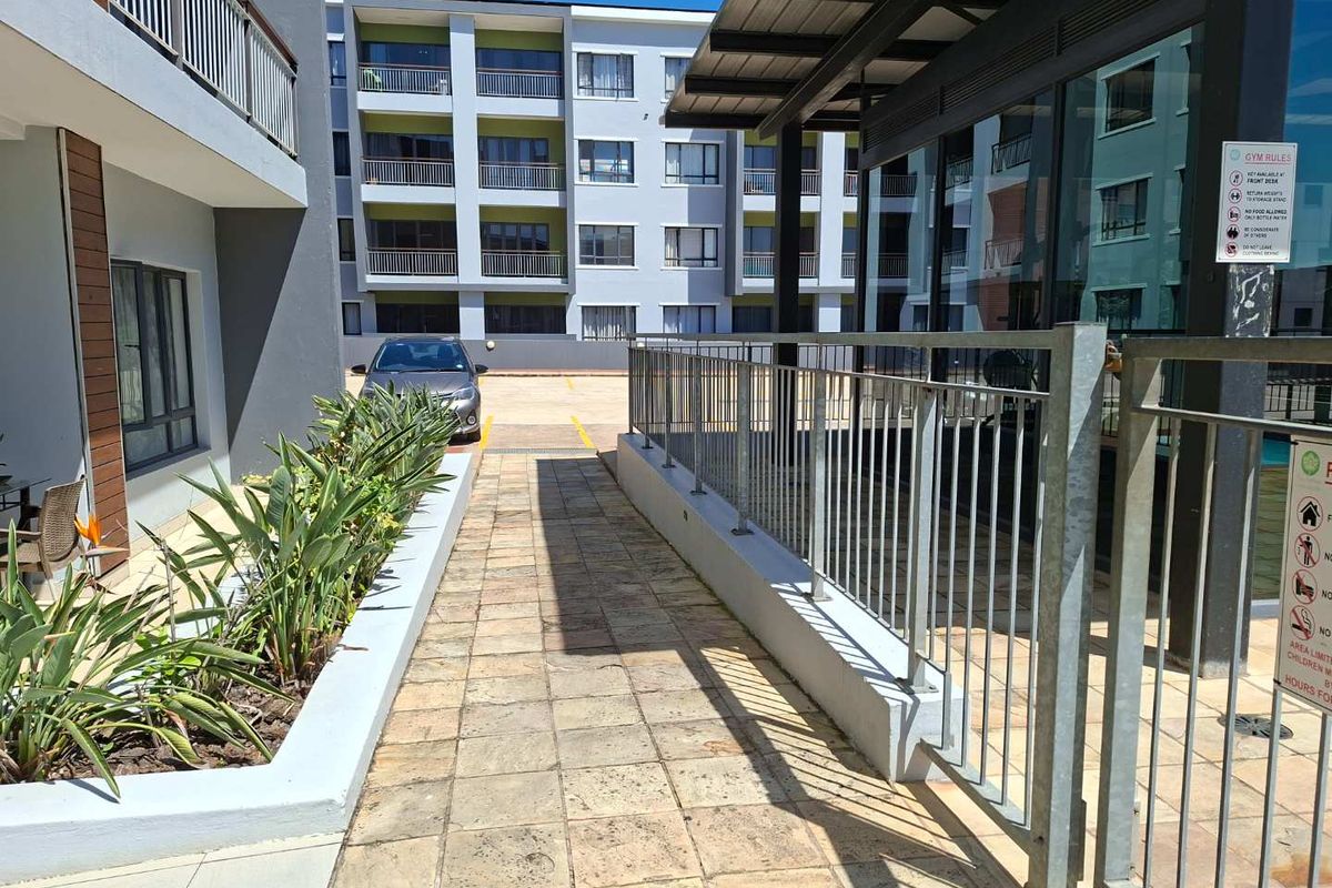 Exterior view of the apartment complex grounds, featuring paved walkways, metal railings, and landscaped garden beds leading toward the residential building.