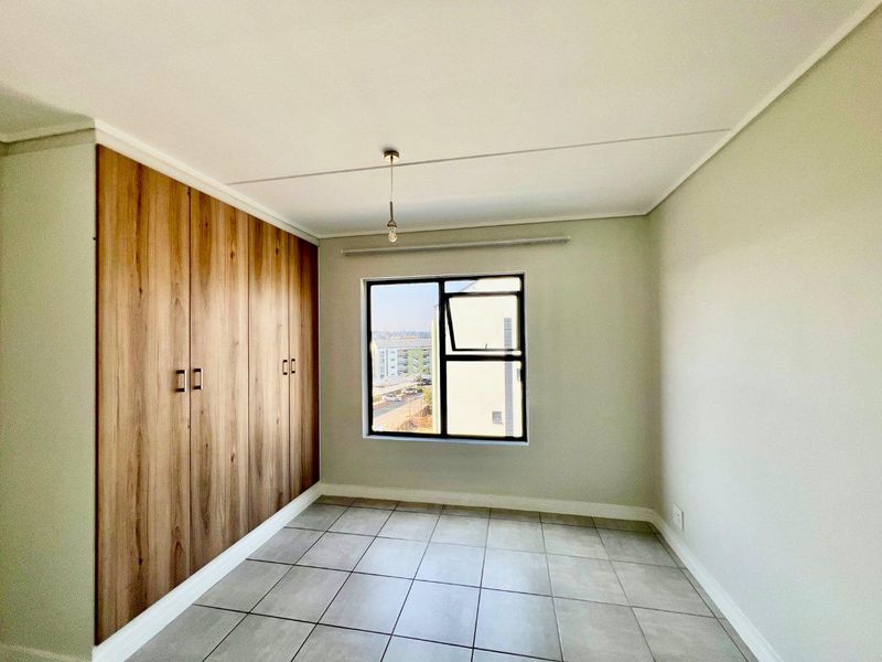 A bright, empty bedroom featuring modern wood-finish built-in cupboards, neutral tiled flooring, and a window providing natural light.