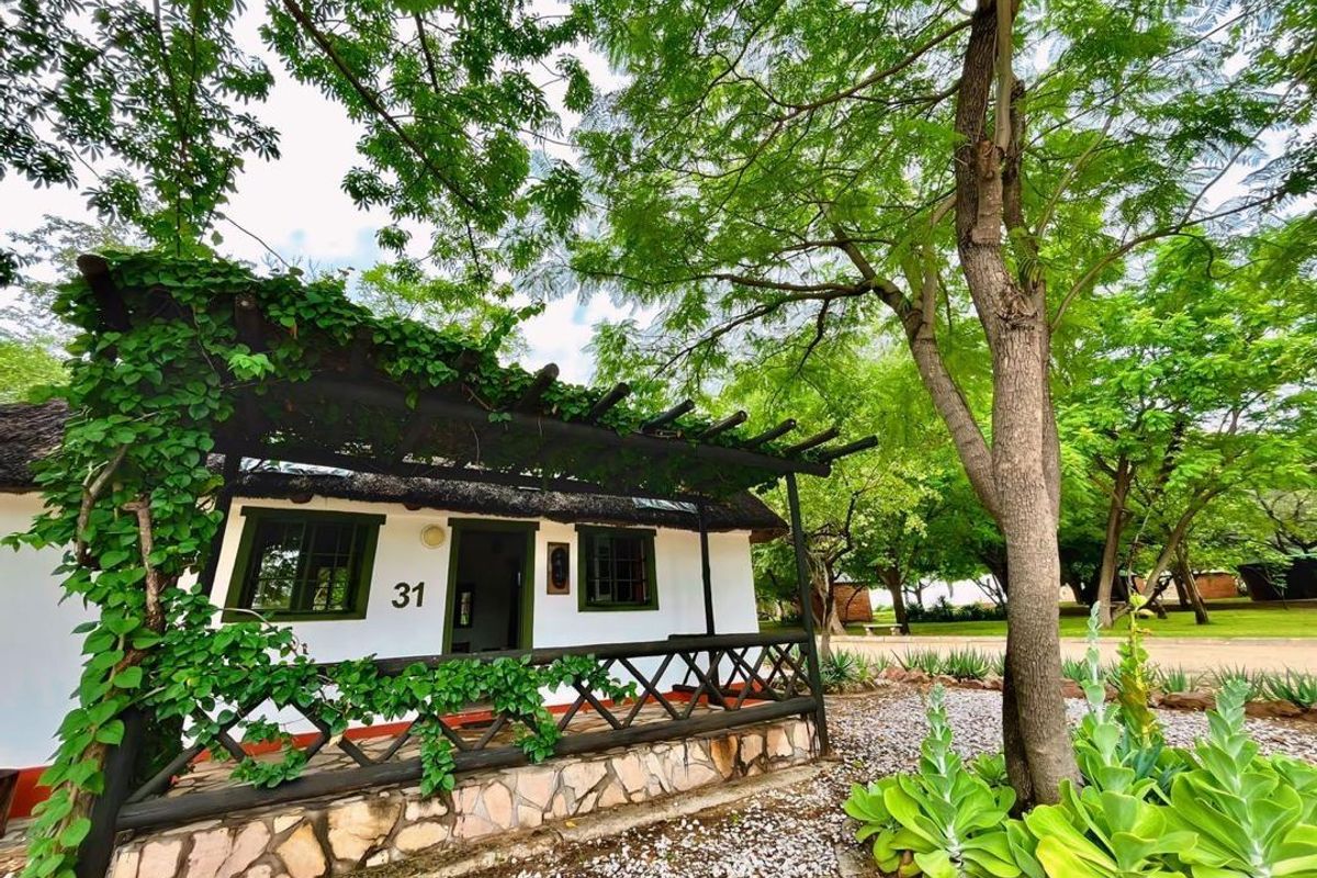 A charming exterior view of a hotel unit featuring a thatched roof, a covered wooden patio, and stone cladding, surrounded by lush garden greenery.