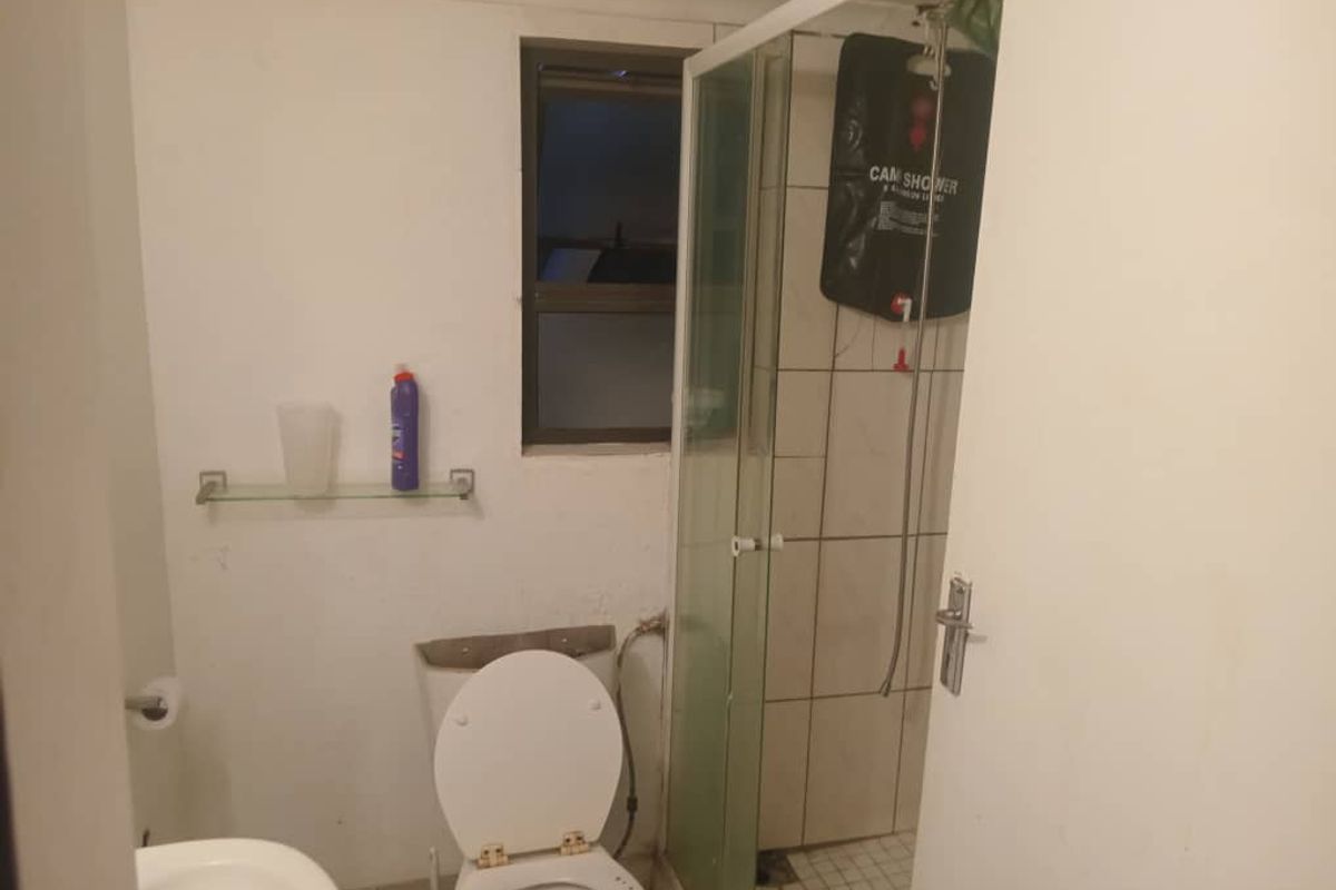 A compact bathroom featuring a shower enclosure, toilet, and basic wall-mounted glass shelf, requiring cosmetic updates and maintenance.