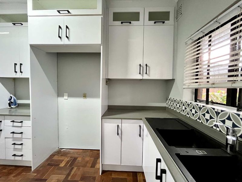 A modern, clean kitchen featuring white cabinetry, grey stone countertops, a double basin sink, and stylish patterned tile backsplash with parquet flooring.