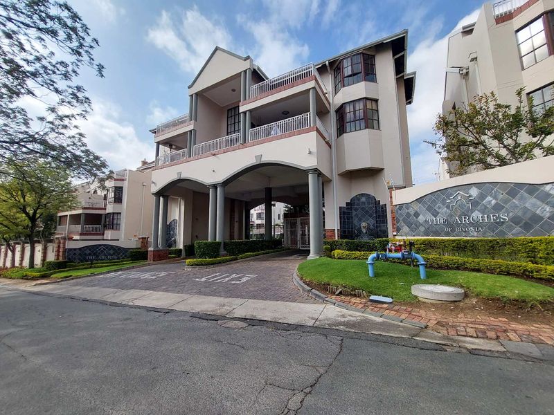 Front elevation of The Arches of Rivonia apartment complex, featuring a paved entrance, stone-clad signage, and multi-level balconies.