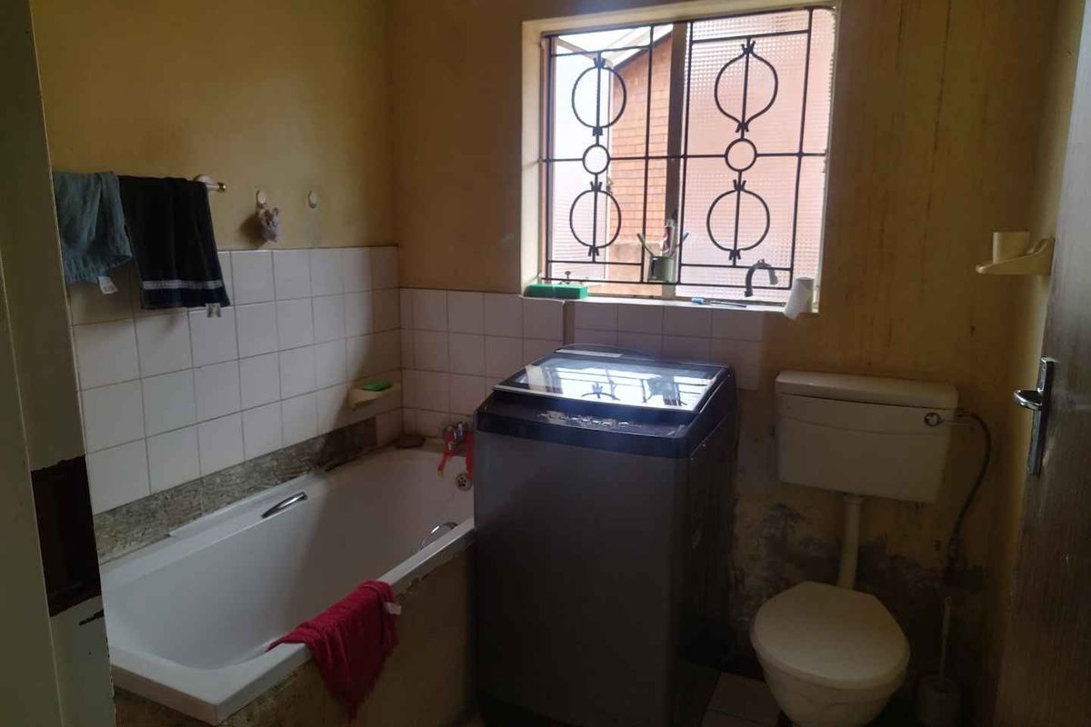A dated bathroom featuring a built-in bathtub, toilet, and window with security bars. The space requires cosmetic updates and wall repairs.