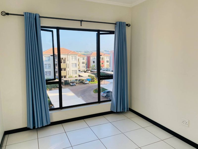 A bright, empty bedroom featuring tiled flooring, large aluminium-framed windows with a neighbourhood view, and neutral wall finishes.