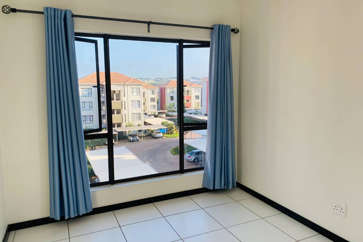 A bright, empty bedroom featuring tiled flooring, large aluminium-framed windows with a neighbourhood view, and neutral wall finishes.
