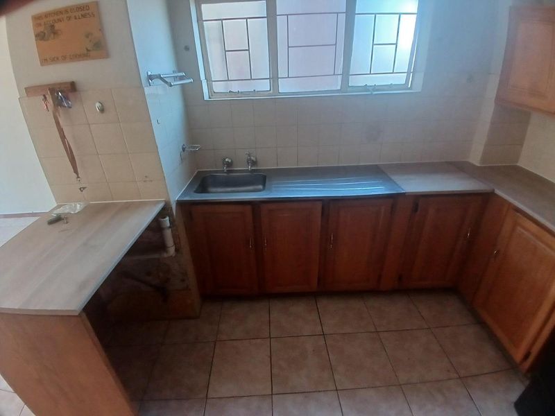A dated kitchen featuring wooden cabinetry, laminate countertops, a stainless steel sink, and tiled flooring, requiring cosmetic updates and repairs to the cabinetry base.