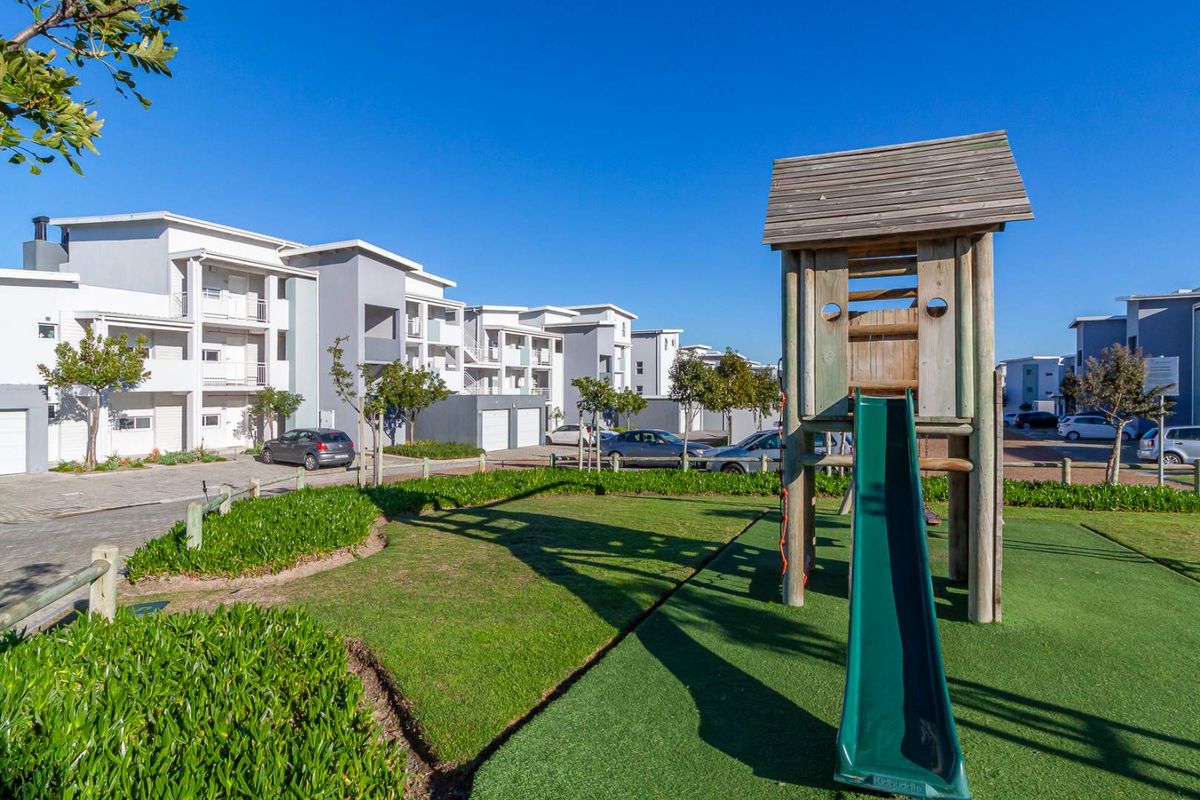 A well-maintained communal garden area featuring a children's playground structure, set within a modern apartment complex with paved parking and landscaped grounds.