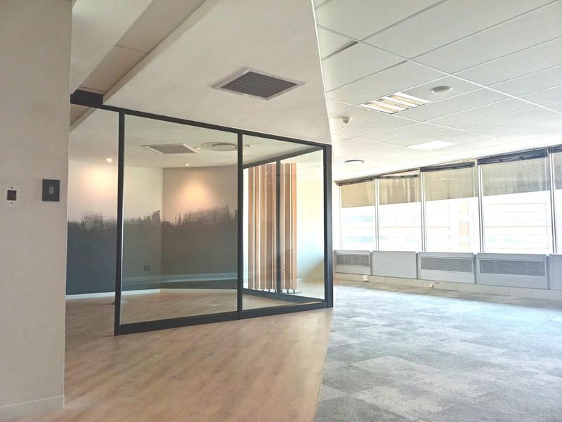 Modern open-plan office space featuring a glass-partitioned meeting room, suspended ceilings, carpet tiles, and large windows providing ample natural light.