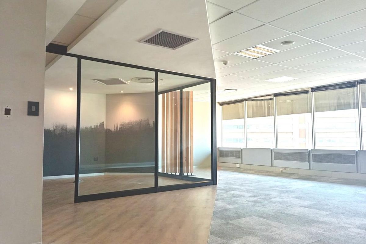 Modern open-plan office space featuring a glass-partitioned meeting room, suspended ceilings, carpet tiles, and large windows providing ample natural light.