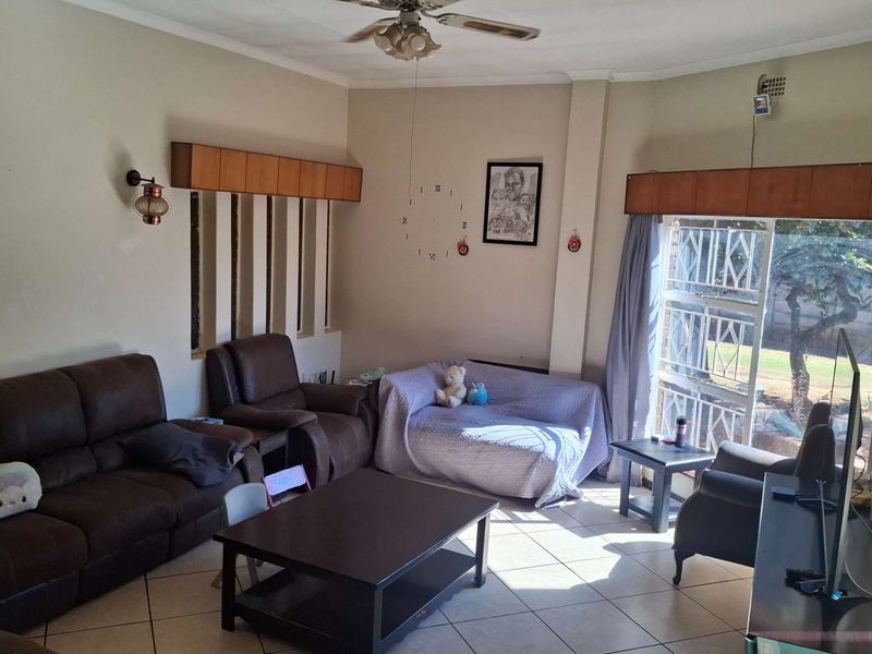 A spacious living room featuring tiled flooring, a ceiling fan, and large windows with security bars and built-in pelmets.