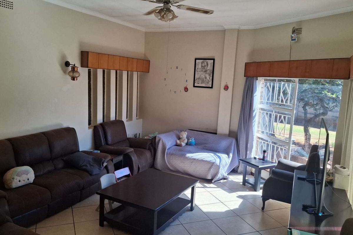 A spacious living room featuring tiled flooring, a ceiling fan, and large windows with security bars and built-in pelmets.