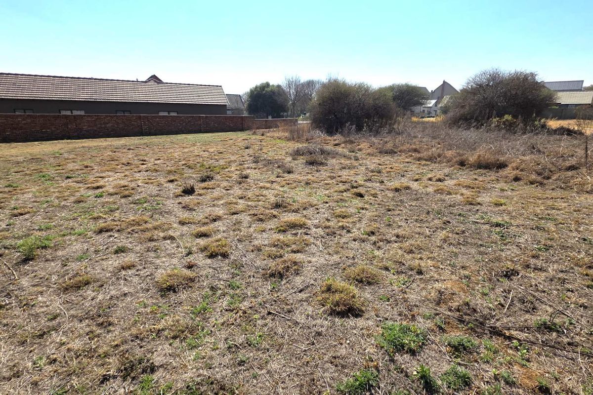 A vacant residential plot featuring dry, unmaintained ground cover and a perimeter brick boundary wall in a suburban setting.
