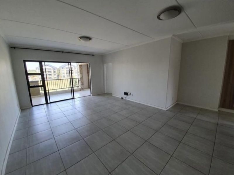 An empty, open-plan living area featuring neutral tiled flooring and a large sliding glass door providing access to an outdoor balcony.