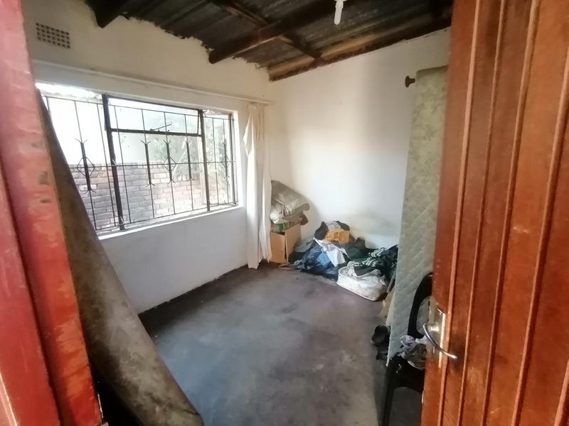 A small bedroom featuring exposed timber rafters, a corrugated metal ceiling, and security bars on the window, requiring significant renovation and cosmetic updates.