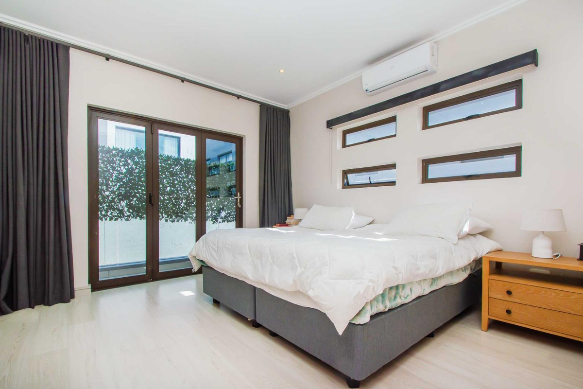 A bright and modern bedroom featuring light laminate flooring, a split-system air conditioner, and horizontal clerestory windows with access to an outdoor area.