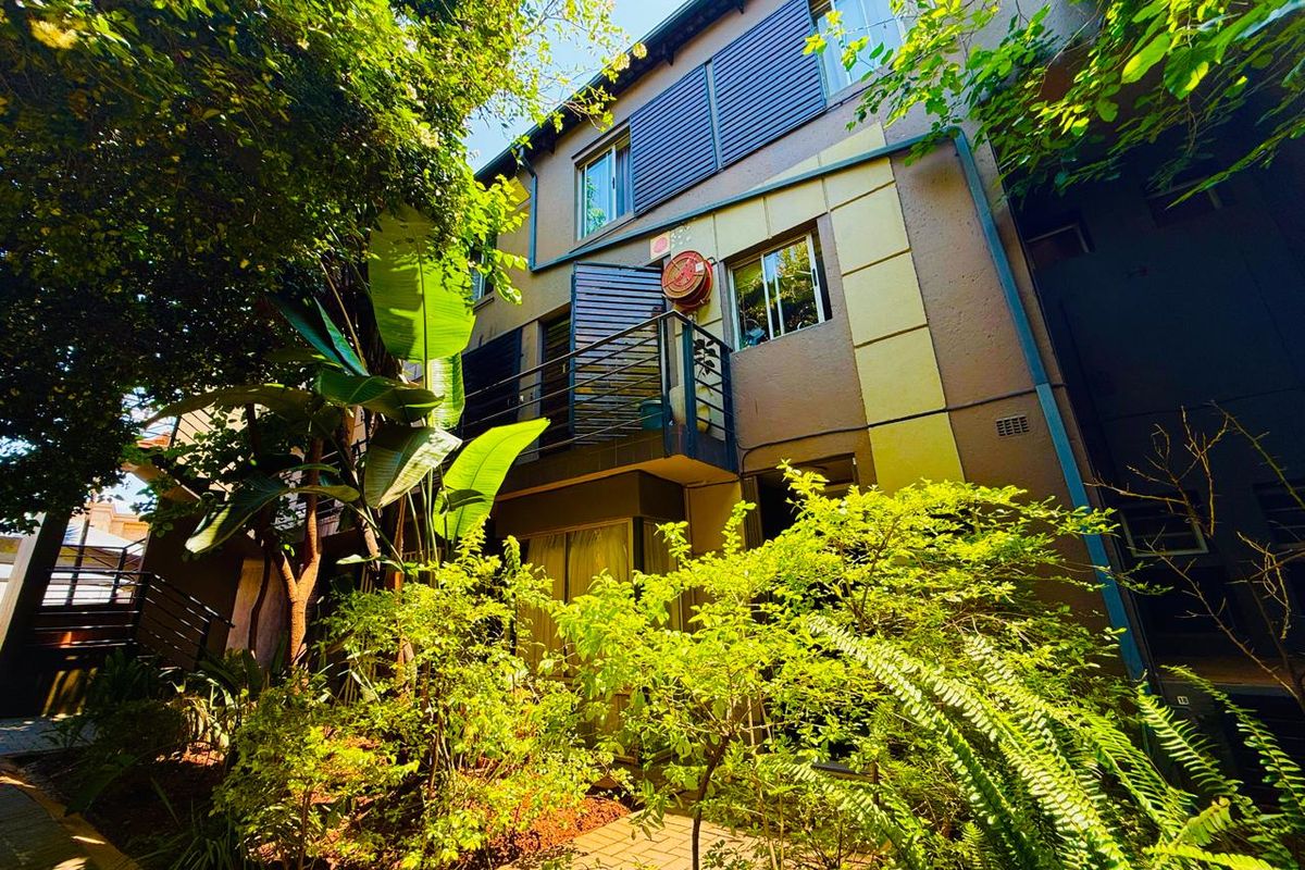 A view of the apartment building exterior featuring a balcony, landscaped garden area, and paved walkway.