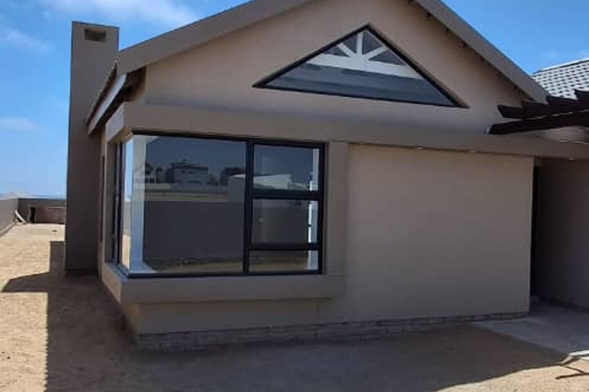 Exterior view of a modern house featuring a pitched roof, triangular gable window, and large corner windows, set on a sandy plot.