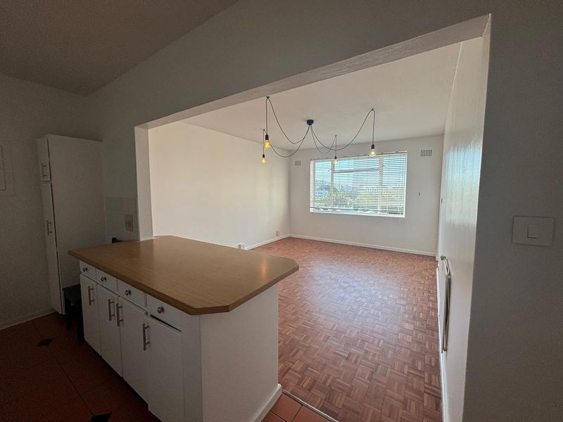 An open-plan living and kitchen area featuring parquet flooring, a breakfast counter, and large windows with venetian blinds.