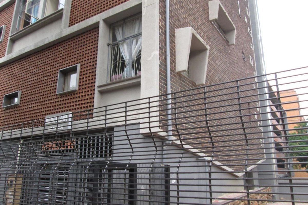 Exterior view of an apartment building facade featuring brickwork, concrete architectural details, and perimeter security fencing.