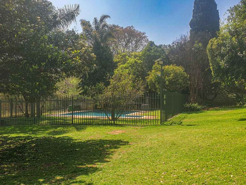 A well-maintained outdoor pool area enclosed by metal security fencing, set within a spacious, lush green garden with mature trees.