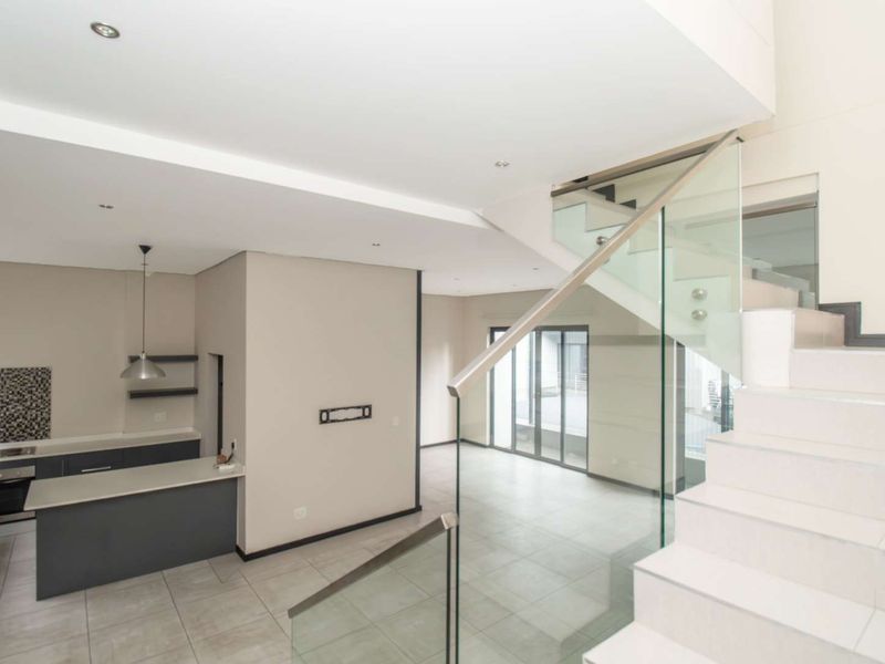 Modern open-plan living area featuring a sleek glass staircase, tiled floors, and a contemporary kitchen with built-in appliances and neutral finishes.