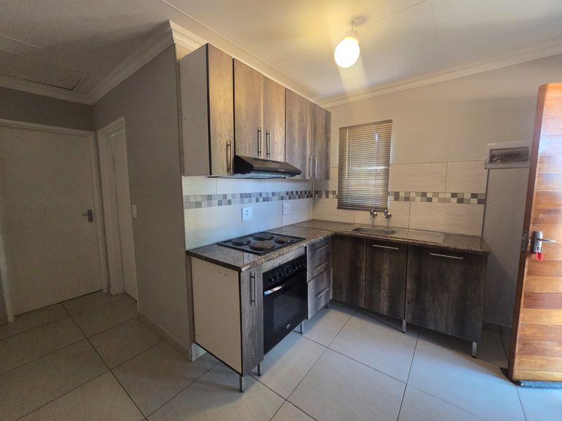 Compact kitchen featuring wood-look cabinetry, built-in electric oven and hob, tiled splashback, and neutral floor tiles.