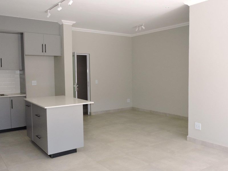 A modern open-plan living and kitchen area featuring grey cabinetry, a central island with stone countertops, tiled flooring, and neutral wall finishes.