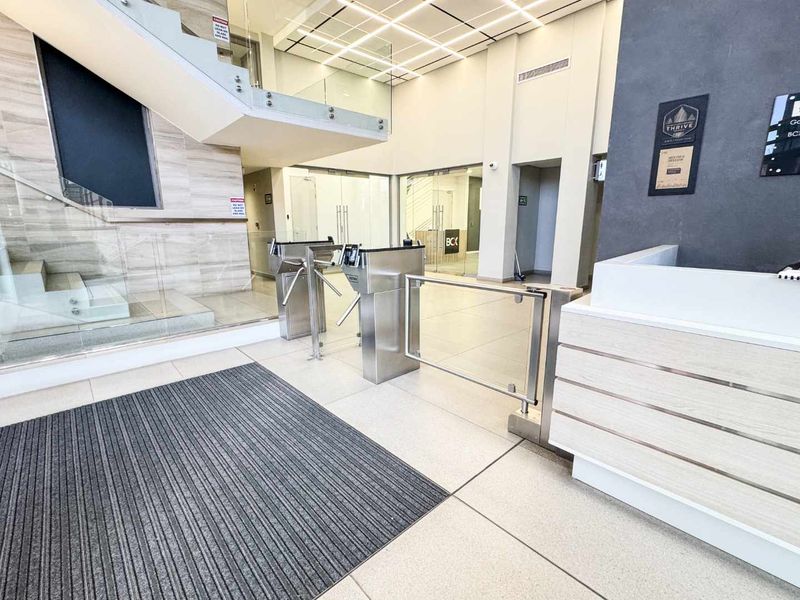 Modern commercial building lobby featuring a reception desk, secure turnstile access gates, and a contemporary glass staircase in a bright, professional environment.