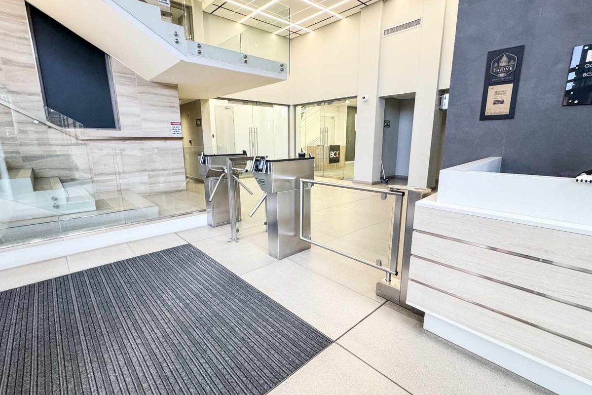 Modern commercial building lobby featuring a reception desk, secure turnstile access gates, and a contemporary glass staircase in a bright, professional environment.