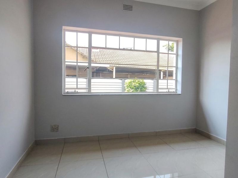 A bright, empty bedroom featuring neutral grey walls, polished floor tiles, and a large window providing natural light.