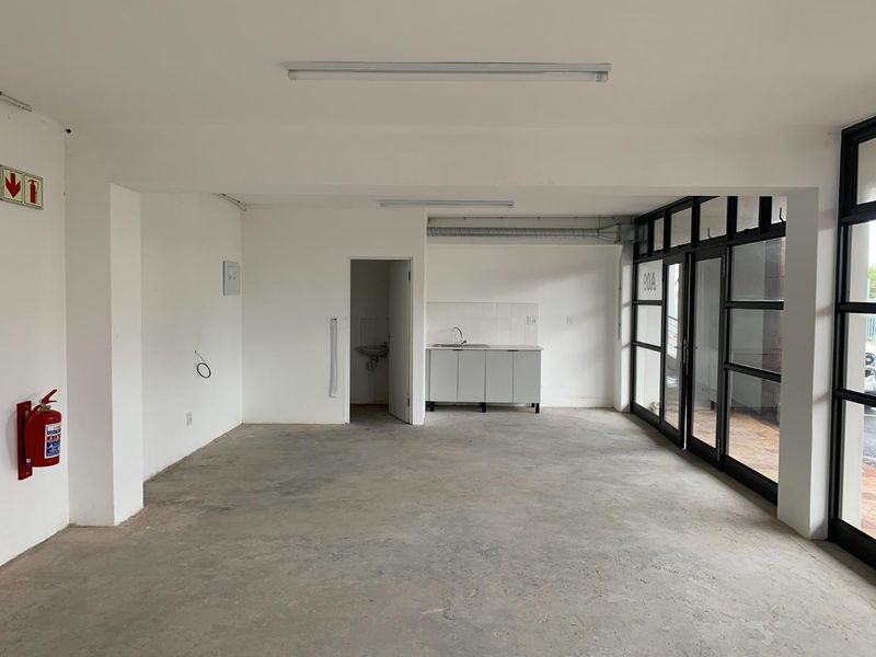 Open-plan commercial or office space featuring a kitchenette, black aluminium framed glass doors, and unfinished concrete flooring, suitable for various business uses.