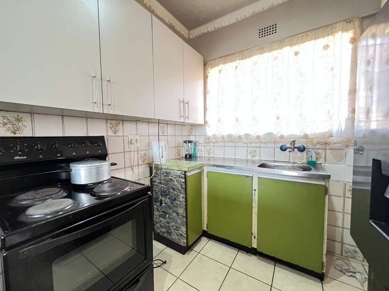 A compact kitchen featuring dated green cabinetry, a stainless steel sink, ceramic tile backsplash, and a freestanding electric stove.