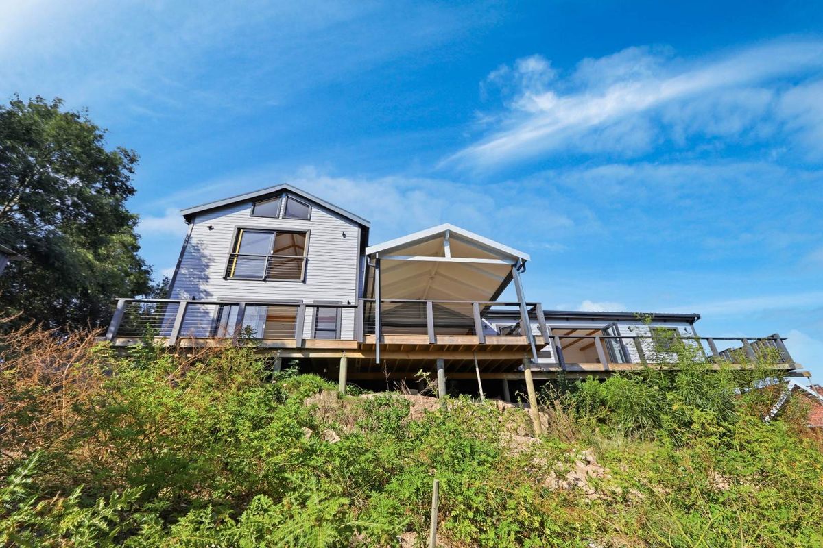 Rear view of a multi-level house featuring a large timber deck, covered patio area, and modern grey cladding set against a natural landscape.