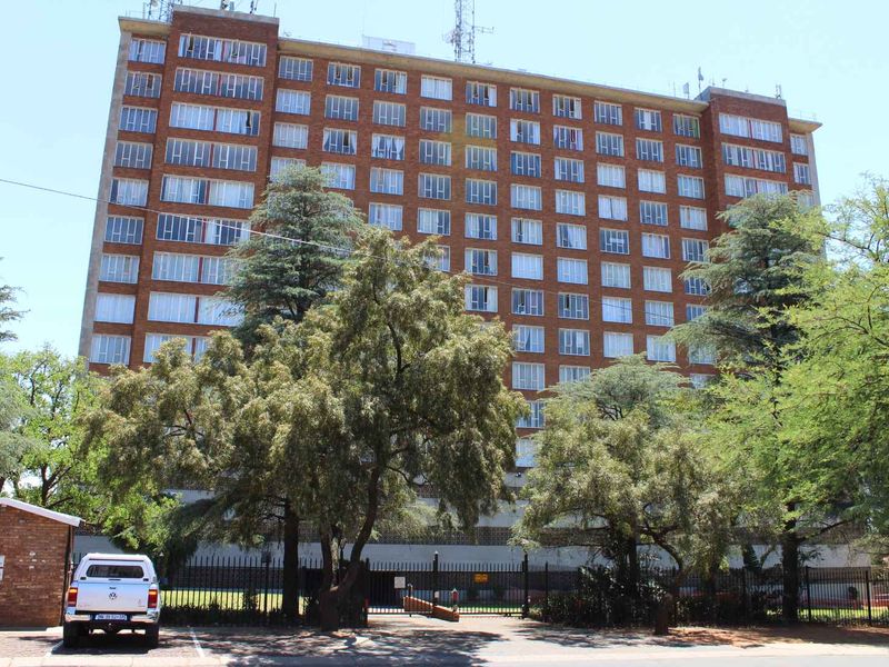 A multi-story brick apartment building viewed from the street, featuring a secure perimeter fence and mature trees in the foreground.