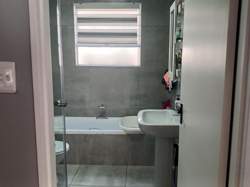 A modern bathroom featuring grey wall and floor tiles, a pedestal basin, and a bathtub with a glass shower screen.