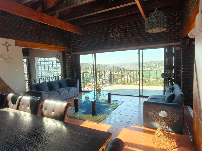 Spacious open-plan living and dining area featuring exposed timber beams, brick walls, and large sliding doors opening onto a balcony with scenic views.