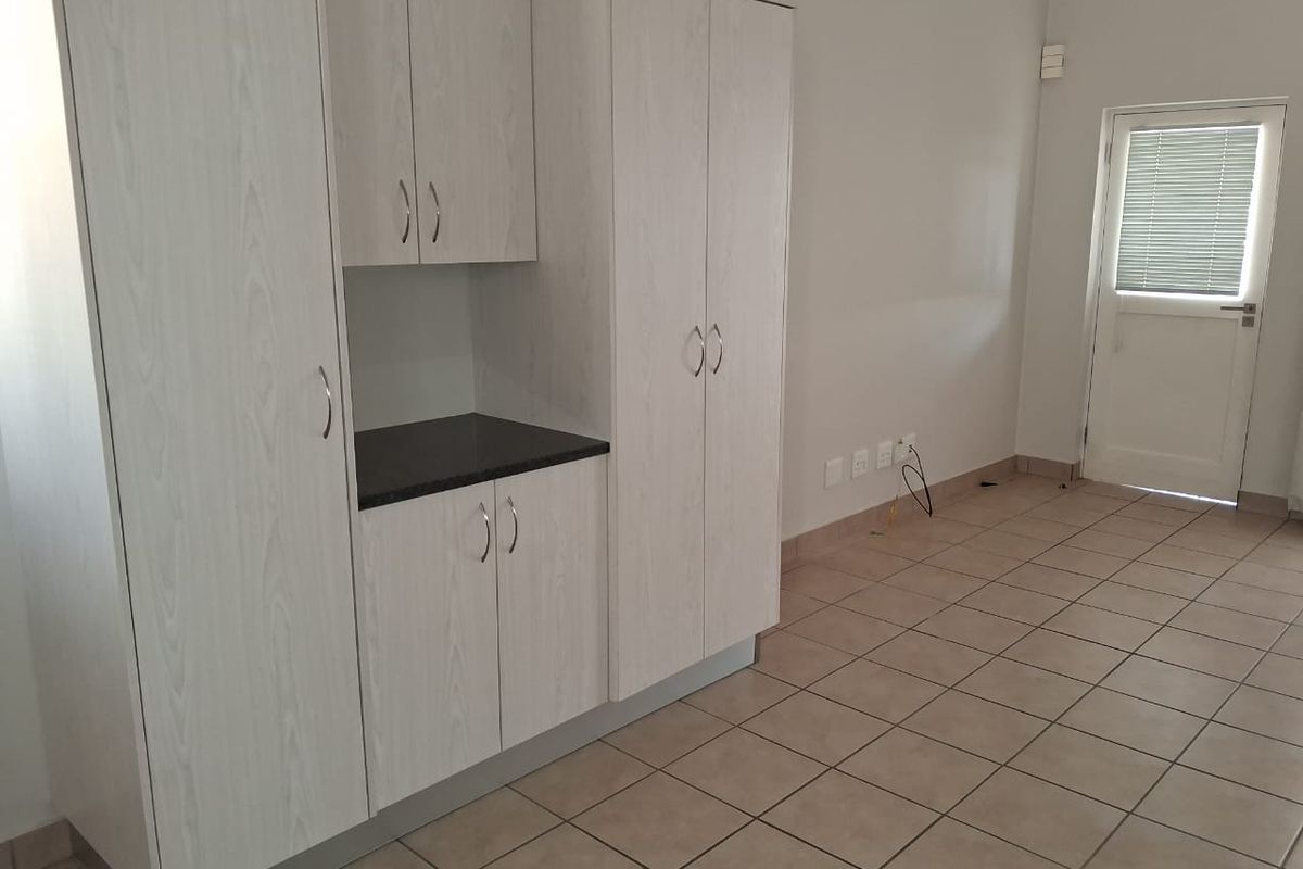 A clean, empty kitchen area featuring light wood-grain built-in cabinetry, dark laminate countertops, and neutral floor tiles, leading to an external door.