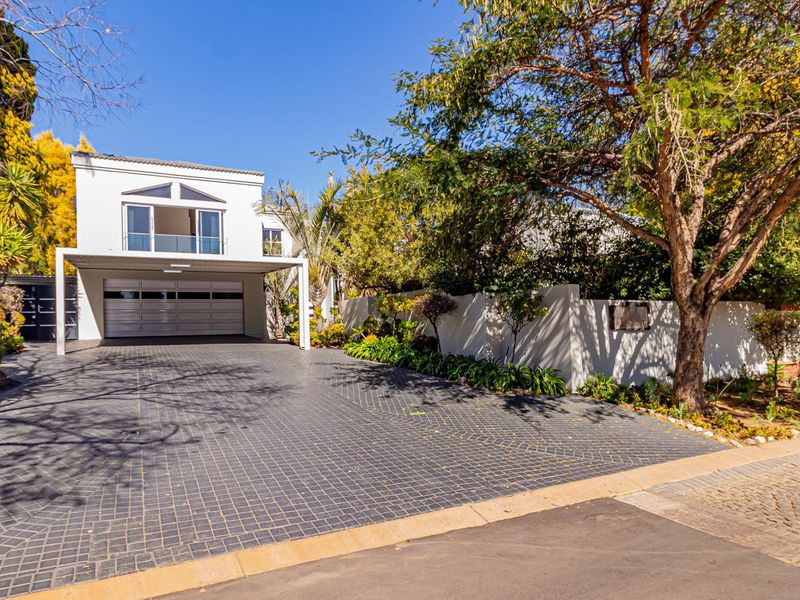 Front elevation of a modern house featuring a spacious paved driveway, double garage, and an upper-level balcony surrounded by established garden landscaping.