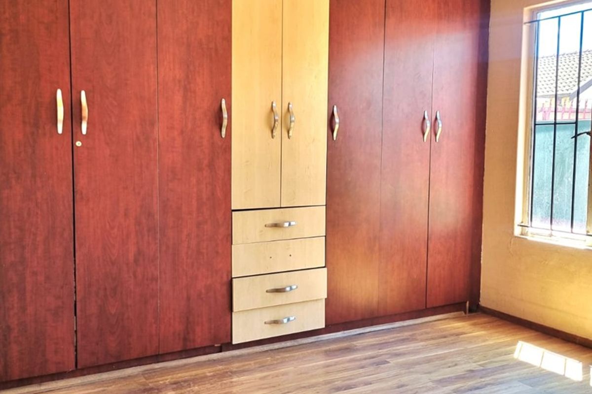 A bright, empty bedroom featuring extensive built-in wooden cupboards, laminate flooring, and security bars on the window.