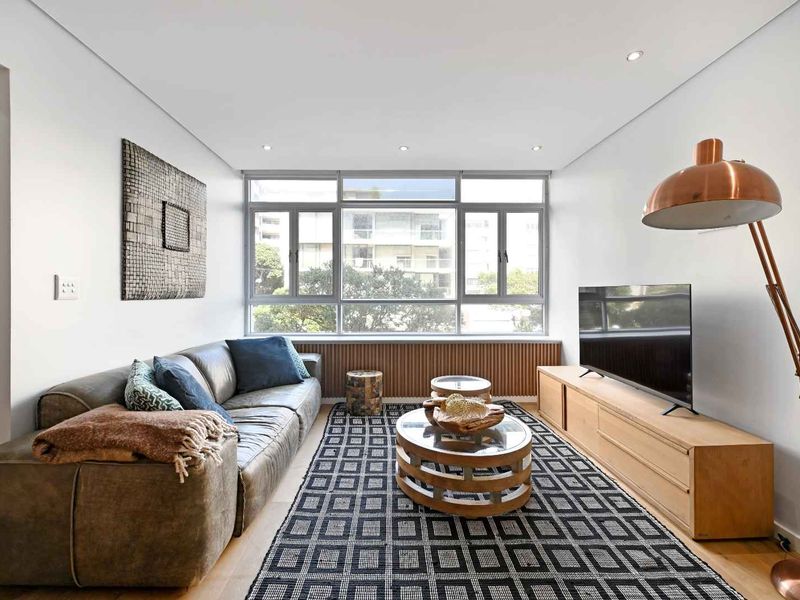 A bright and modern living room featuring hardwood flooring, recessed downlighting, and a large window with timber framing and a decorative radiator cover.