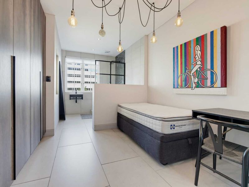 Modern bedroom featuring built-in cupboards, tiled flooring, and an open-plan en-suite bathroom with a glass shower partition and wall-mounted basin.