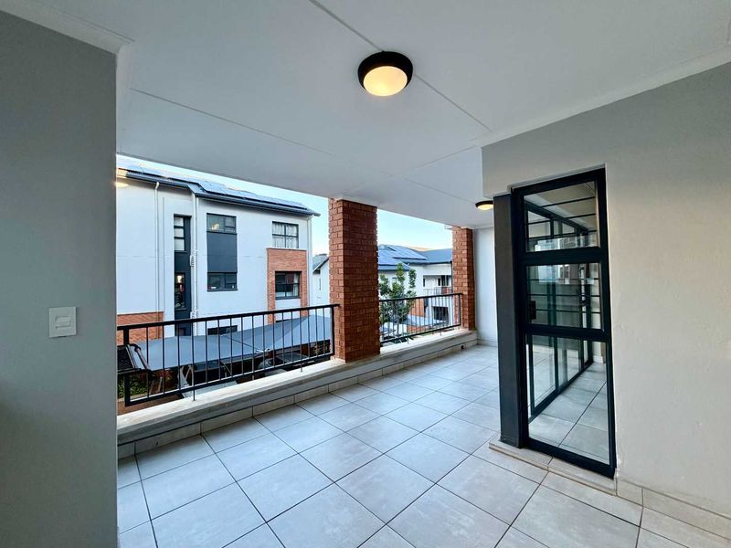 A clean, covered balcony featuring tiled flooring, brick support pillars, and a modern metal balustrade, offering a view of the surrounding apartment complex.