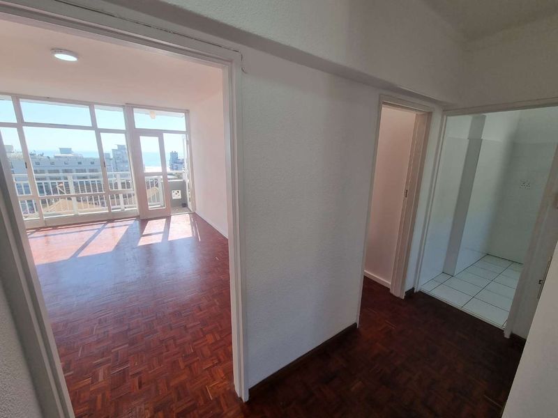 A bright, empty hallway featuring classic parquet flooring, leading into a sunlit living area with large windows and a separate tiled utility space.