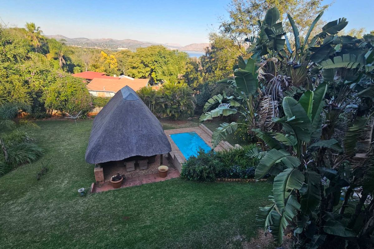 Elevated view of a lush garden featuring a swimming pool and a thatched-roof lapa, set against a scenic landscape backdrop.